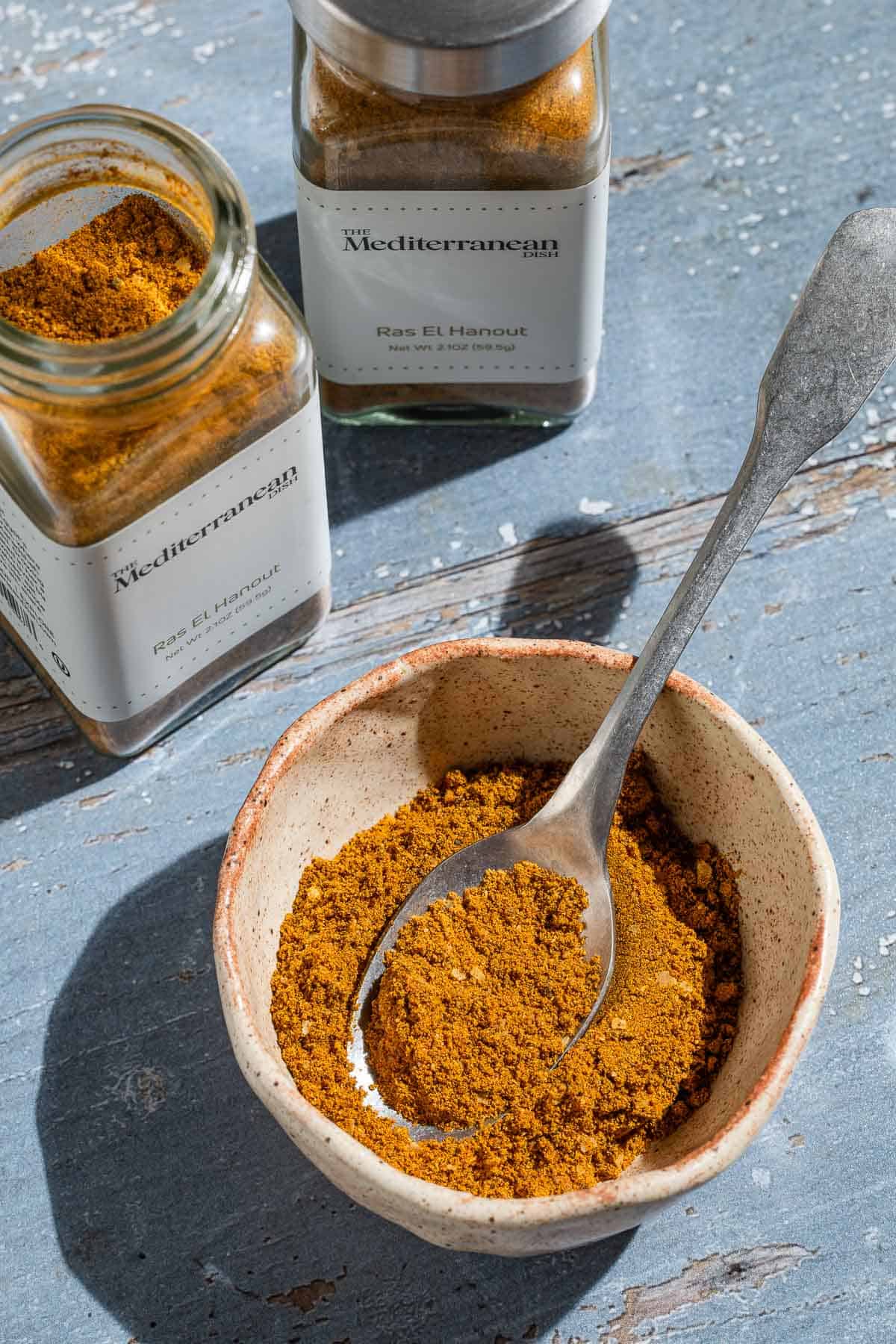 Ras el hanout in a bowl with a spoon. Next to this are two jars of ras el hanout from the mediterranean dish.