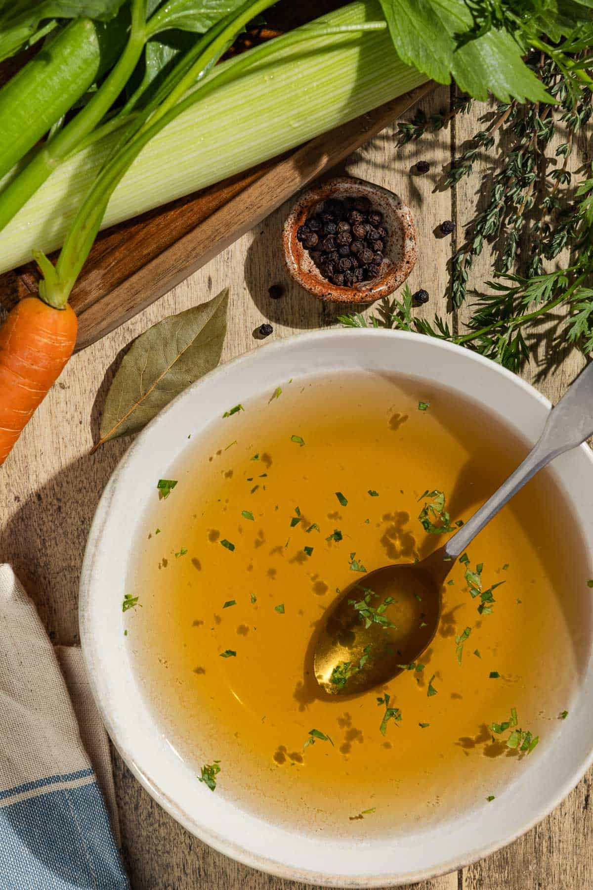 An overhead photo of homemade vegetable stock in a bowl with a spoon surrounded by a leek, carrot, celery, fresh herbs, and a small bowl of black peppercorns.