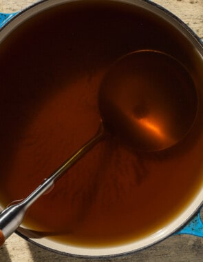 Homemade vegetable stock in a pot with a ladle. This is surrounded by carrots, a lee, celery, a bay leaf, thyme and a small bowl of black peppercorns.