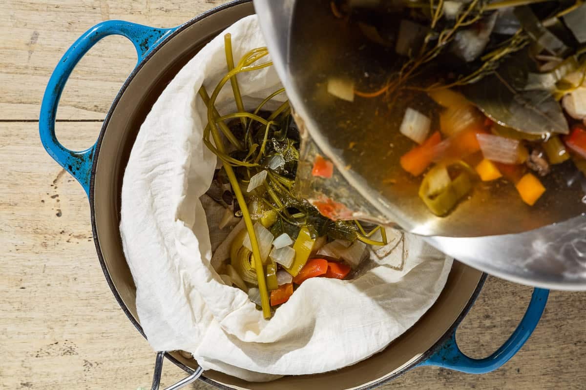 The homemade vegetable stock being strained through a mesh strainer lined with cheesecloth set over a pot.