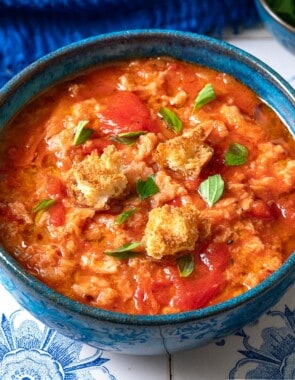 A close up of one bowl of pappa al pomodoro garnished with pieces of ciabatta bread and basil. Next to this are small bowls of olive oil, basil, and ciabatta bread pieces.