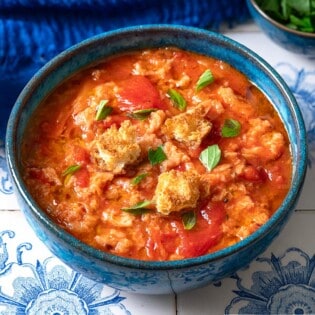 A close up of one bowl of pappa al pomodoro garnished with pieces of ciabatta bread and basil. Next to this are small bowls of olive oil, basil, and ciabatta bread pieces.