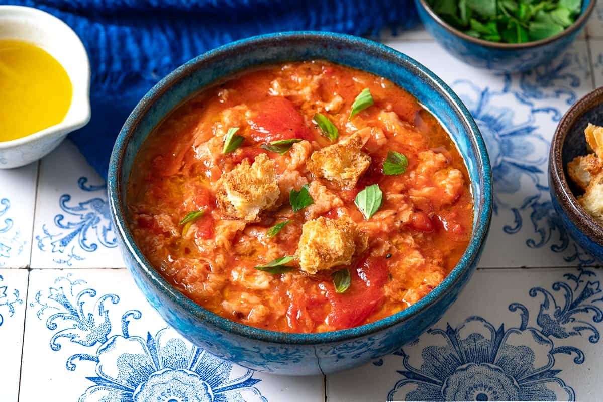 A close up of one bowl of pappa al pomodoro garnished with pieces of ciabatta bread and basil. Next to this are small bowls of olive oil, basil, and ciabatta bread pieces.