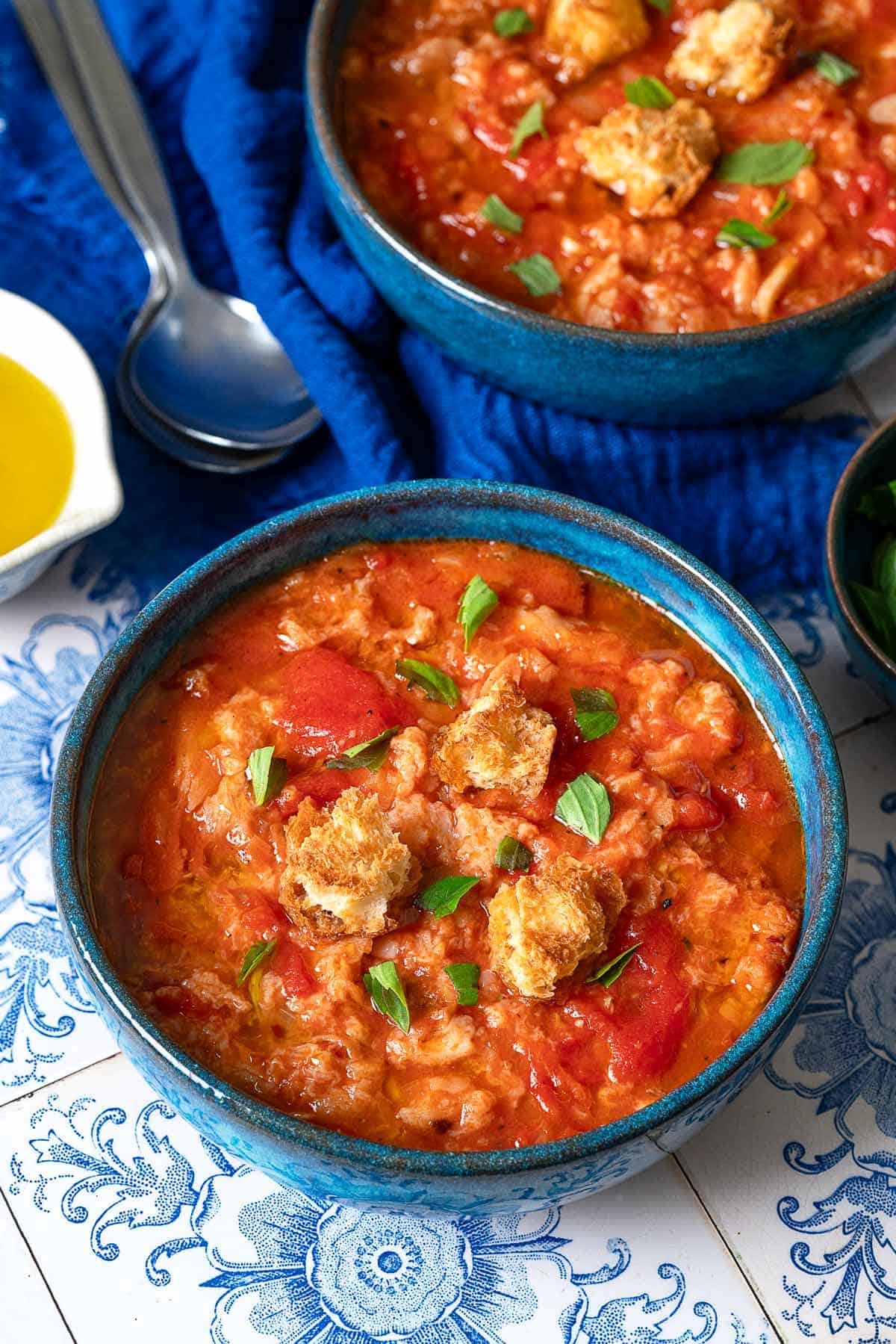 Two bowls of pappa al pomodoro garnished with pieces of ciabatta bread and basil next to two spoons, a blue towel and a bowl of olive oil.