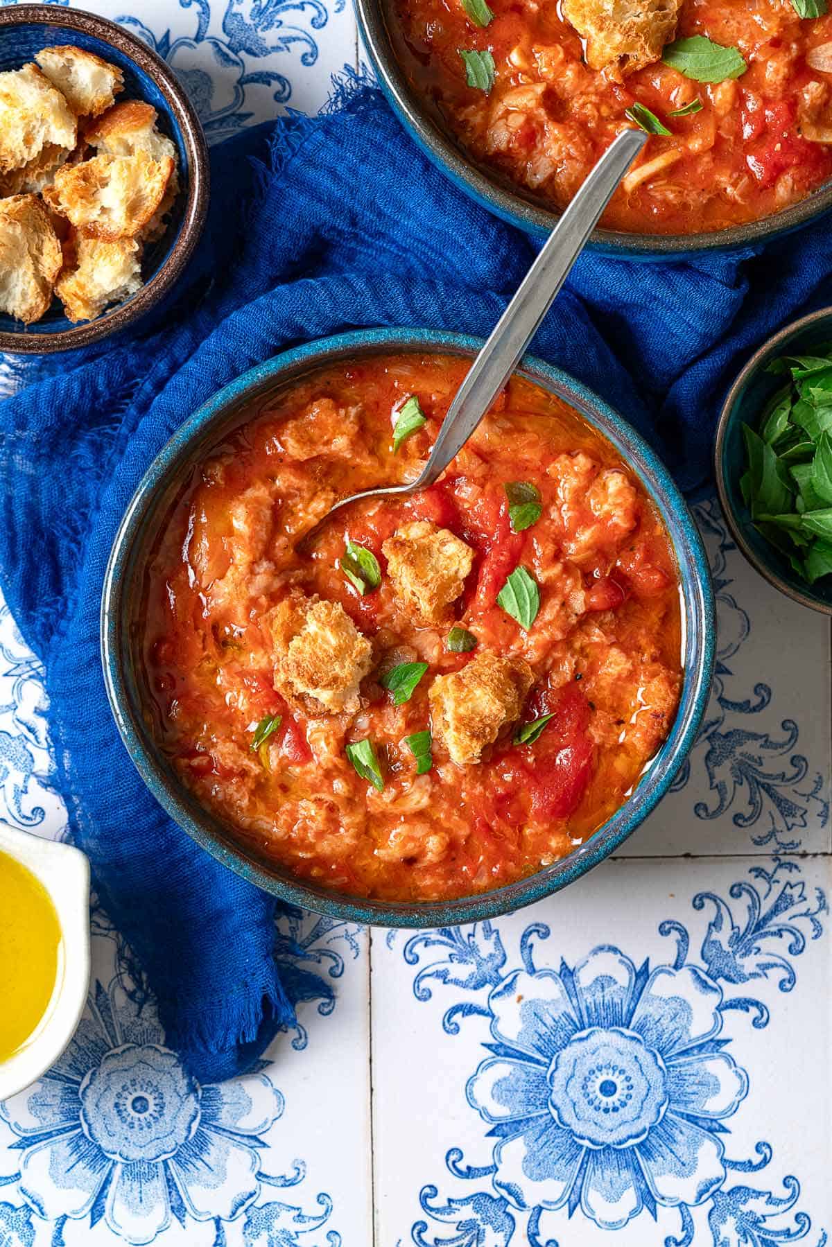 An overhead photo of two bowls of pappa al pomodoro garnished with pieces of ciabatta bread and basil, one with a spoon. Next to these are small bowls of olive oil, basil, more ciabatta pieces and a blue towel.