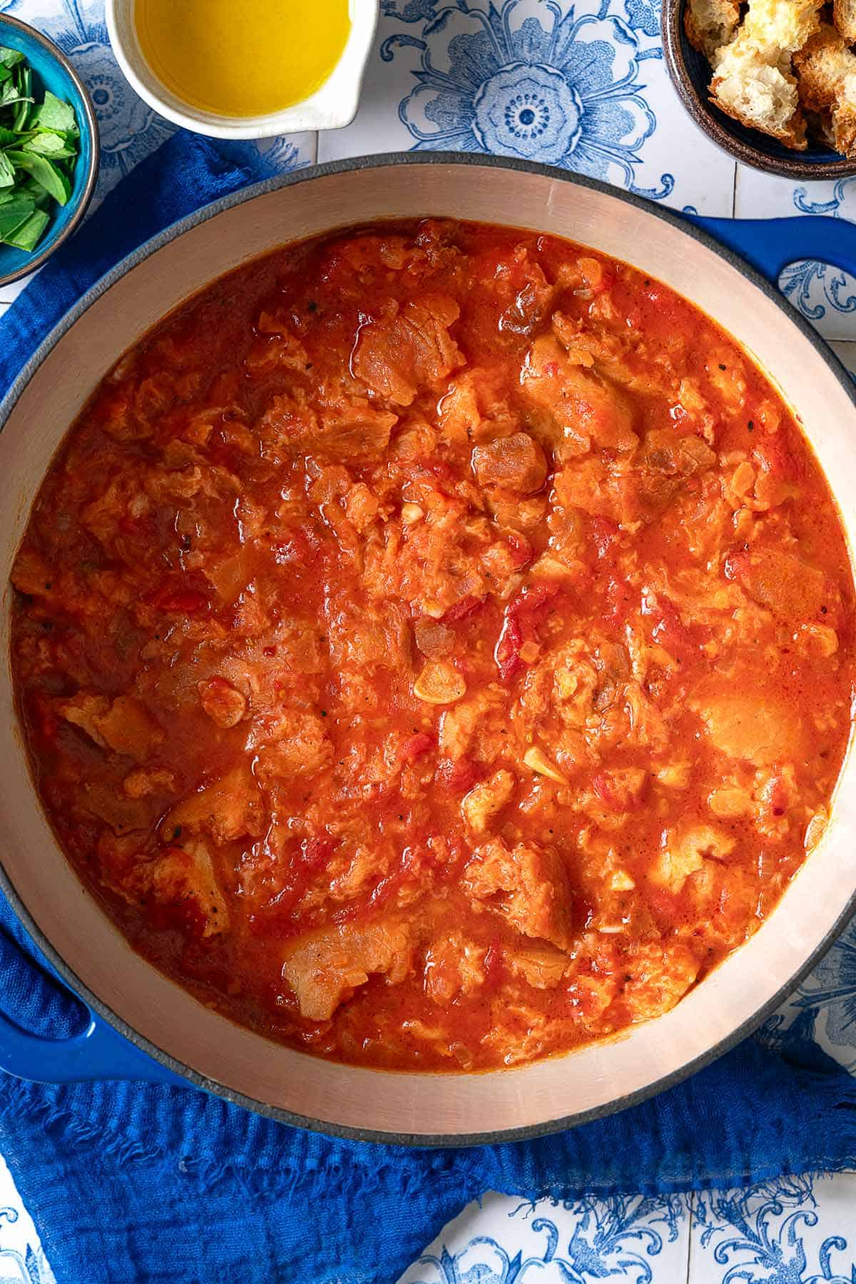 An overhead photo of pappa al pomodoro in a large pot on a blue towel. Next to this are small bowls of basil, olive oil and pieces of ciabatta bread.