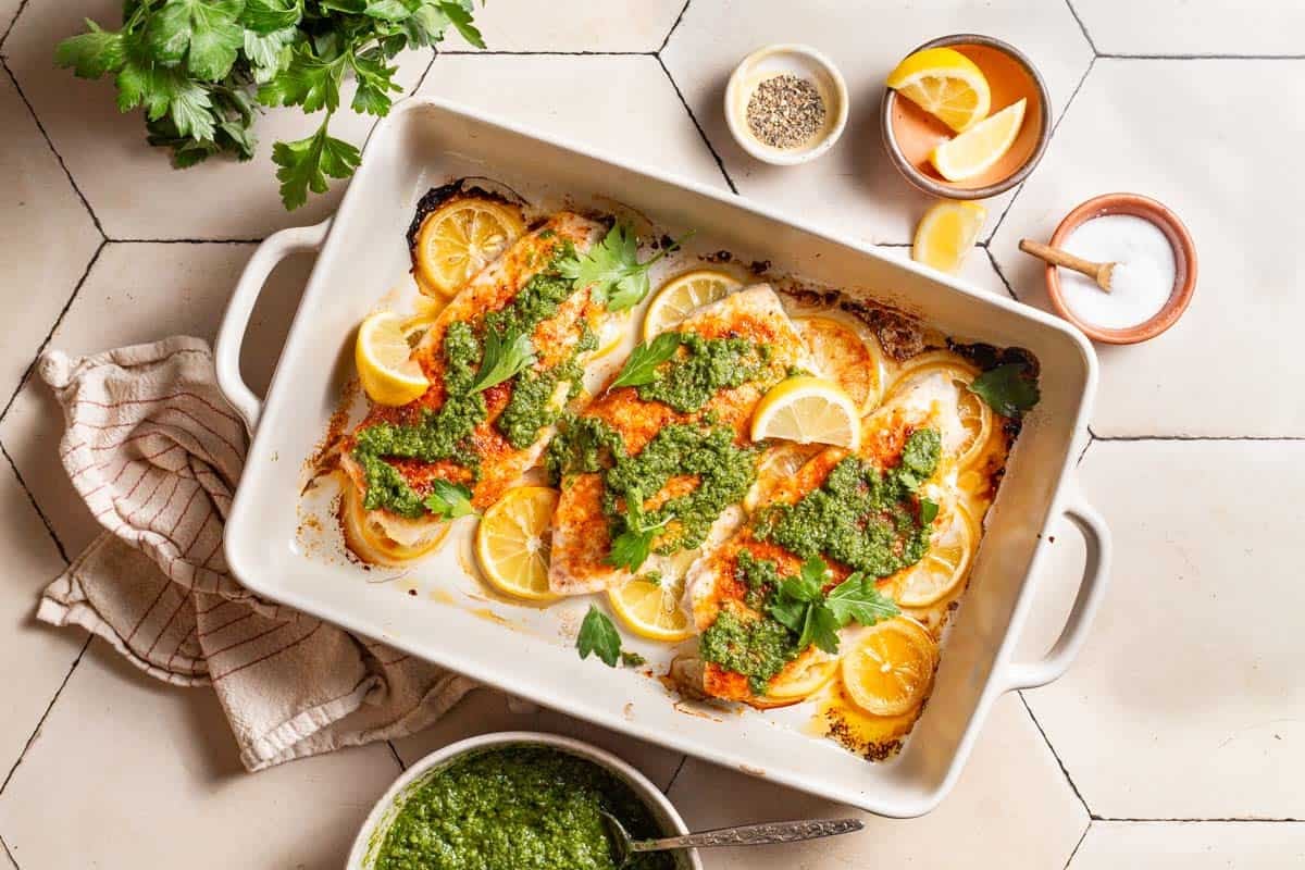 A close up of 3 baked red snapper fillets topped with zhoug and parsley laying on lemon rounds in a baking dish. Surrounding this is a bunch of parsley, a cloth napkin, and bowls of salt, pepper, zhoug and lemon slices.