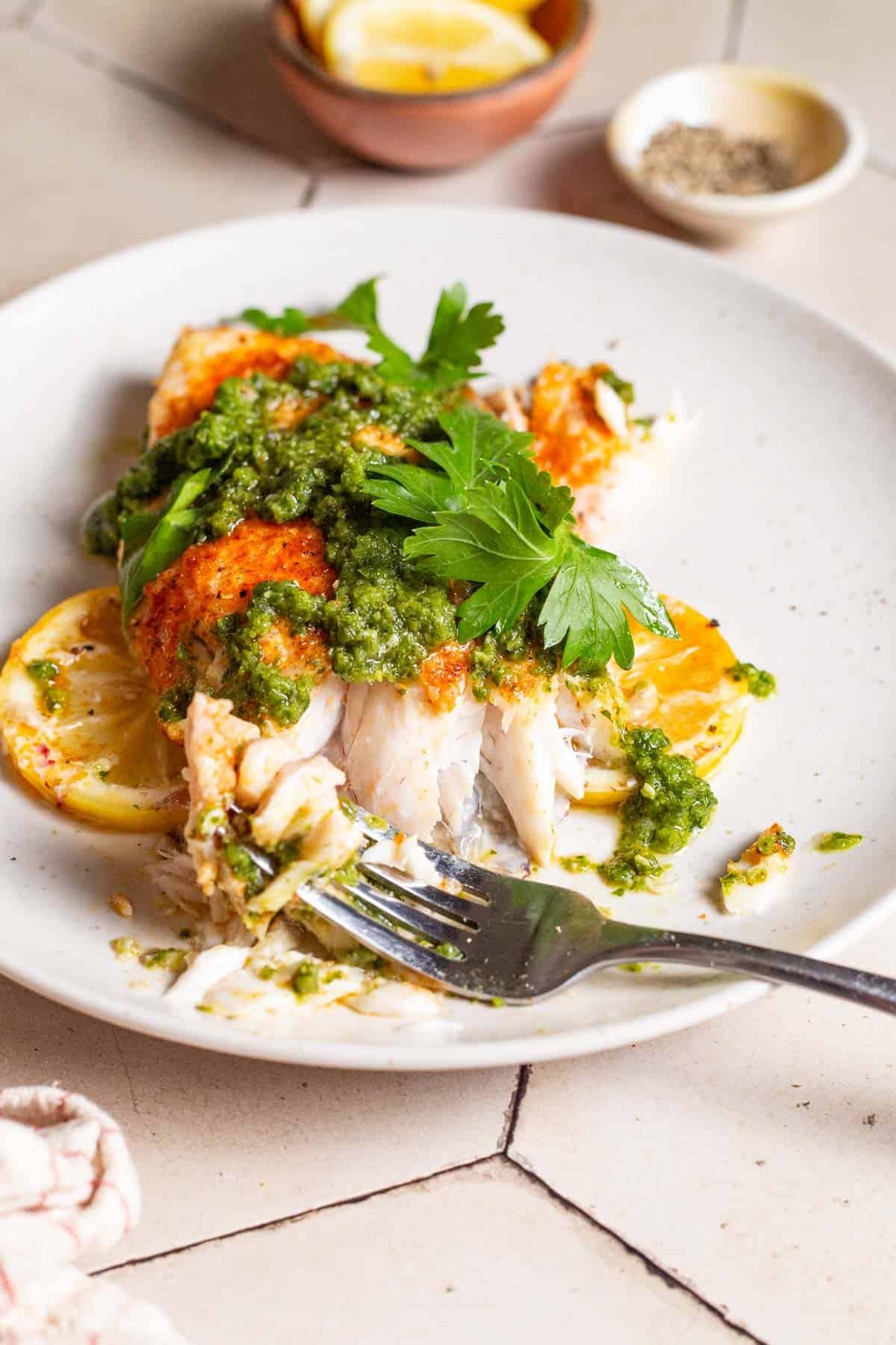 A close up of a baked snapper fillet topped with zhoug and parsley with some lemon slices on a plate with a fork. Next this is a cloth napkin, and small bowls of pepper and lemon slices.