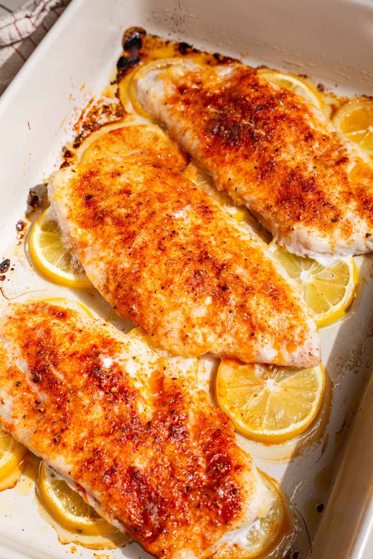 A close up of 3 baked red snapper fillets laying on lemon rounds in a baking dish.