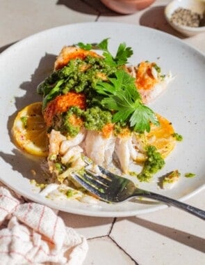 A close up of a baked snapper fillet topped with zhoug and parsley with some lemon slices on a plate with a fork. Next this is a cloth napkin and small bowls of pepper and lemon slices.