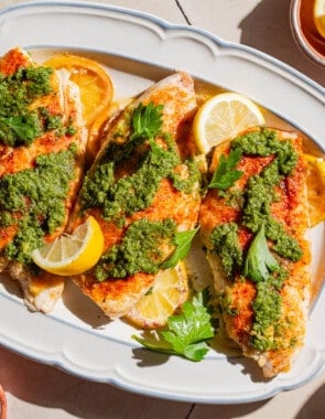 An overhead photo of 3 baked red snapper fillets topped with zhoug and parsley laying on lemon rounds on a serving platter. Surrounding this is a bunch of parsley, a cloth napkin, and bowls of salt, pepper, zhoug and lemon slices.
