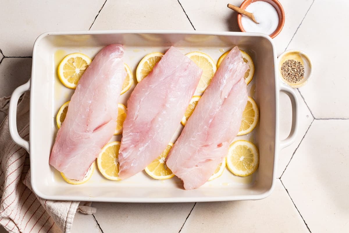 3 uncooked, unseasoned red snapper fillets laying on lemon rounds in a baking dish. Next to this are small bowls of salt and pepper and a cloth napkin.