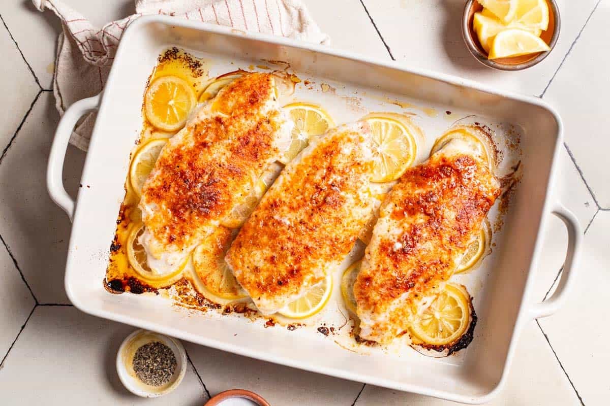 An overhead photo of 3 baked red snapper fillets laying on lemon rounds in a baking dish. Next to this are small bowls of salt, pepper and lemon slices and a cloth napkin.