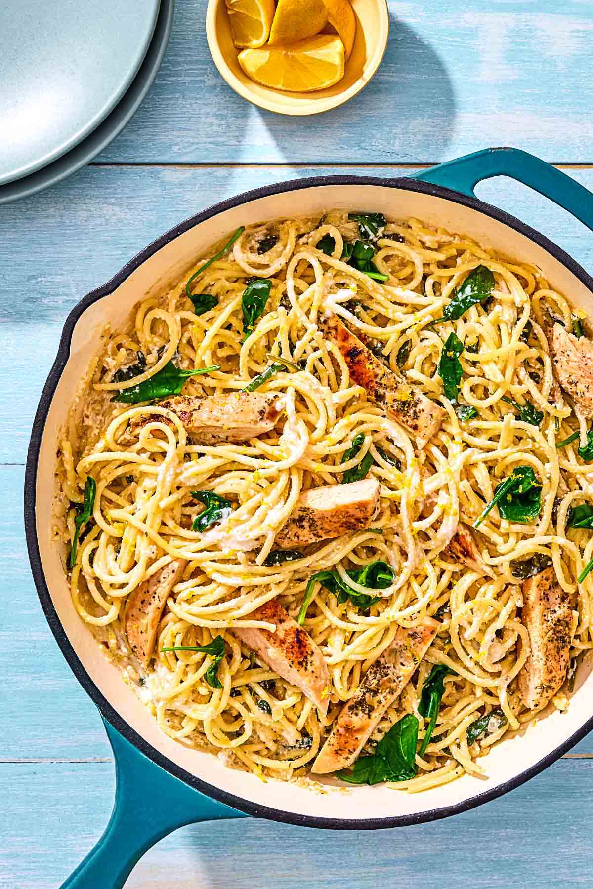 An overhead photo of lemon chicken pasta in a skillet next to a bowl of lemon wedges and a stack of 2 plates.