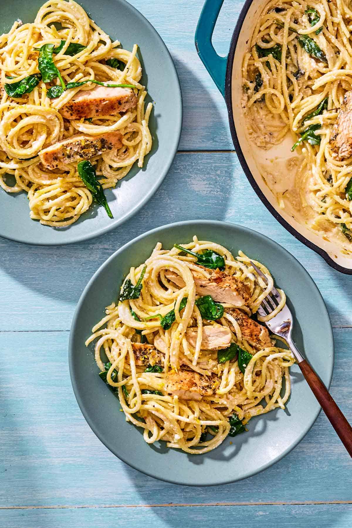 An overhead photo of 2 plates of lemon chicken pasta, one with a fork. Next these is the skillet with the rest of the pasta.