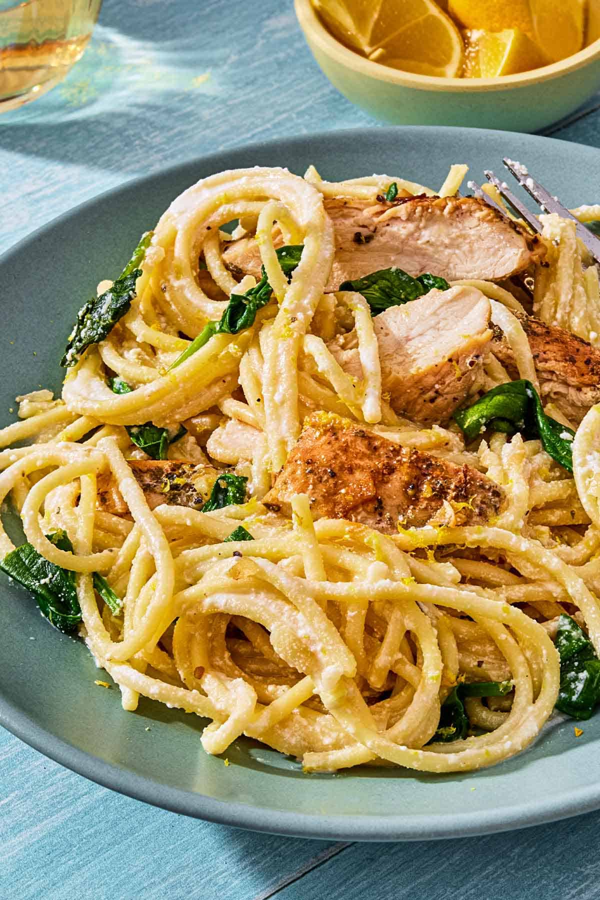 A close up of lemon chicken pasta on a plate with a fork next to a bowl of lemon wedges.