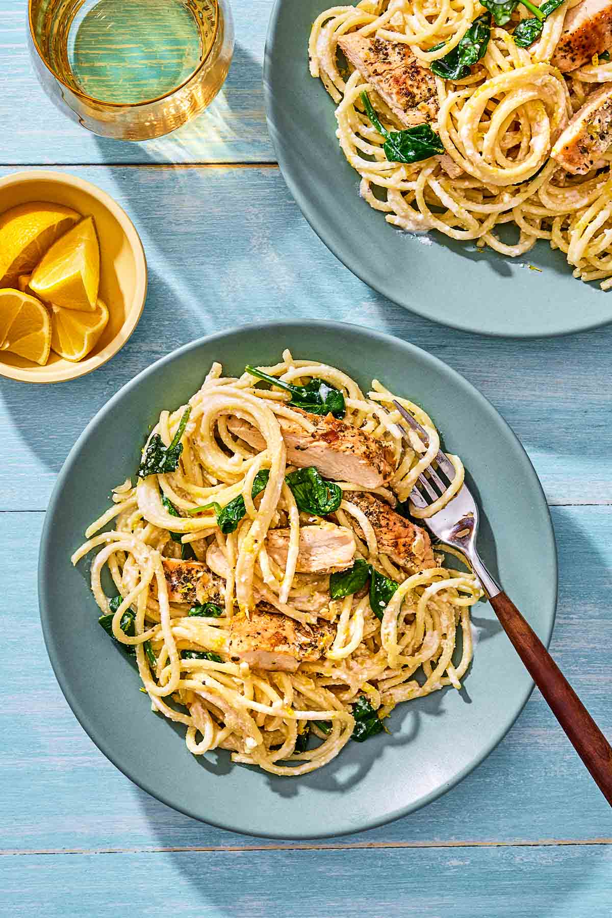 Two plates of lemon chicken pasta beside the skillet and a bowl of lemon wedges.