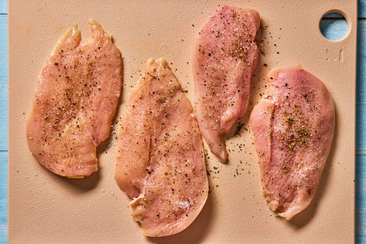 4 uncooked chicken breast fillets seasoned with salt and pepper on a cutting board.