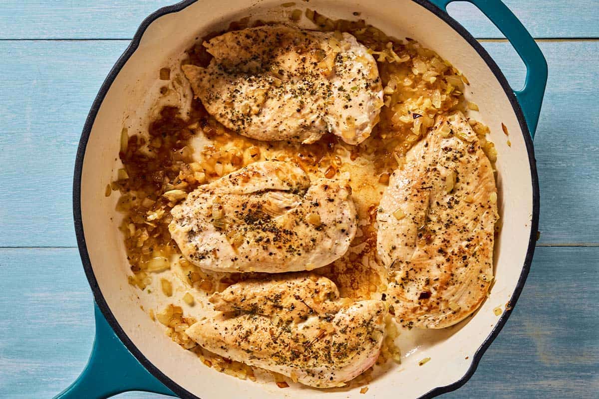 4 chicken breast fillets seasoned with salt and pepper being cooked with onions and garlic in a skillet.