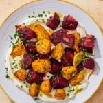 Roasted beets on a layer of labneh spread on a plate and topped with chives and a drizzle of apple cider vinaigrette next to a serving spoon.