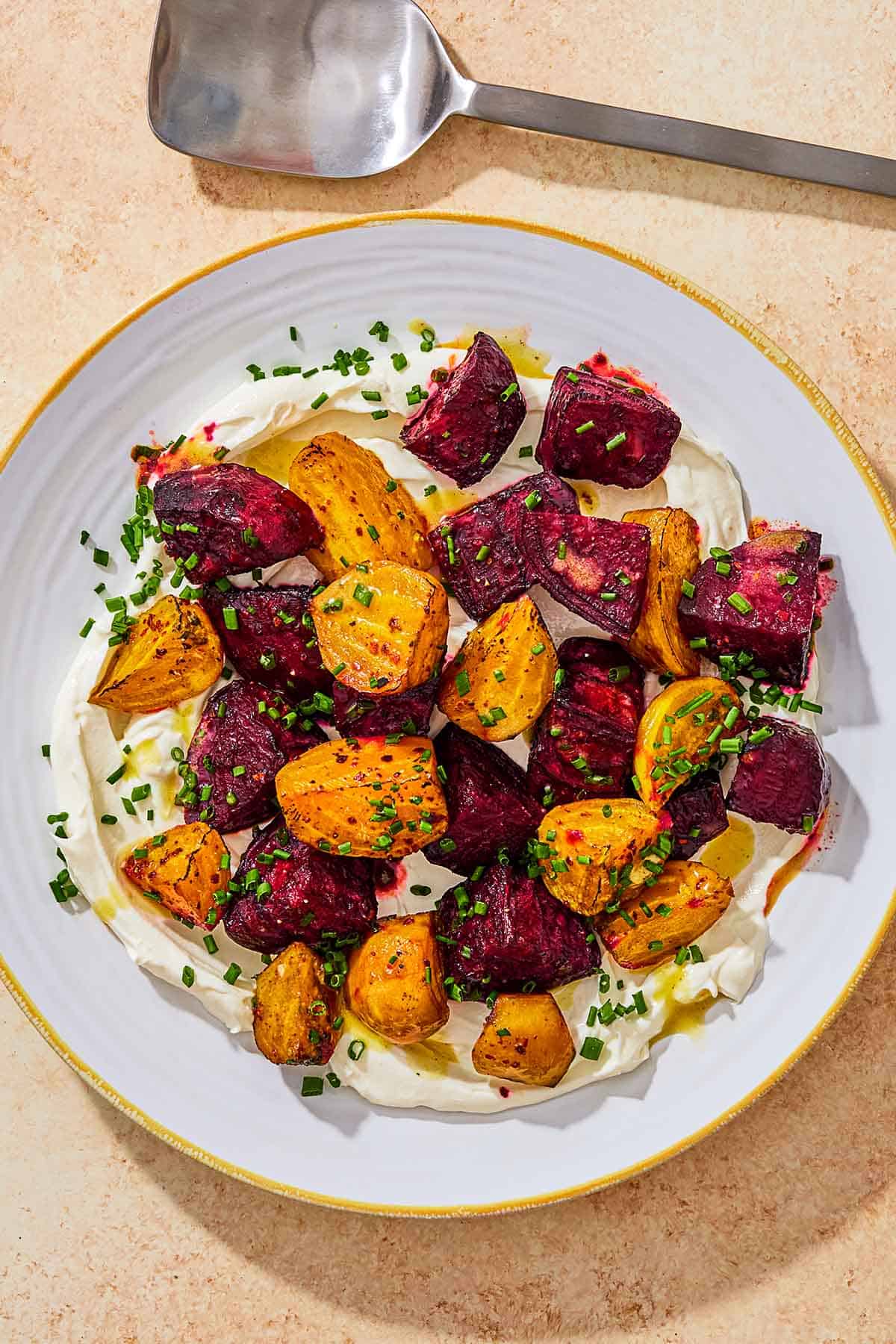 Roasted beets on a layer of labneh spread on a plate and topped with chives and a drizzle of apple cider vinaigrette next to a serving spoon.