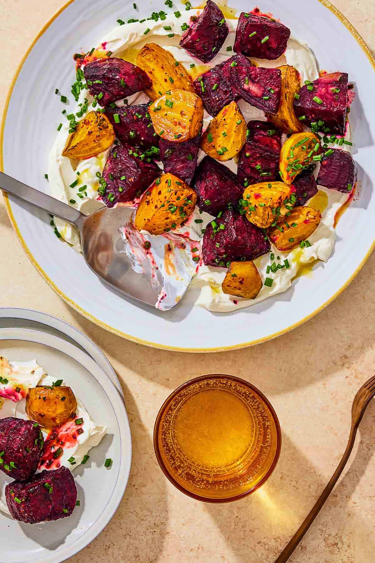 An overhead photo of roasted beets on a layer of labneh spread on a platter and topped with chives and a drizzle of apple cider vinaigrette with a serving spoon. Next to this is a serving of the roasted beets on a plate, a glass of water, and a fork.