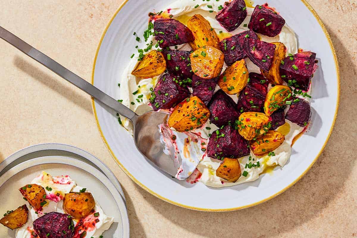 An overhead photo of roasted beets on a layer of labneh spread on a platter and topped with chives and a drizzle of apple cider vinaigrette with a serving spoon. Next to this is a serving of the roasted beets on a plate.
