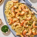 An overhead photo of shrimp cacio e pepe on a serving platter with serving utensils. Next to this is small bowls of pecorino romano cheese and parsley.