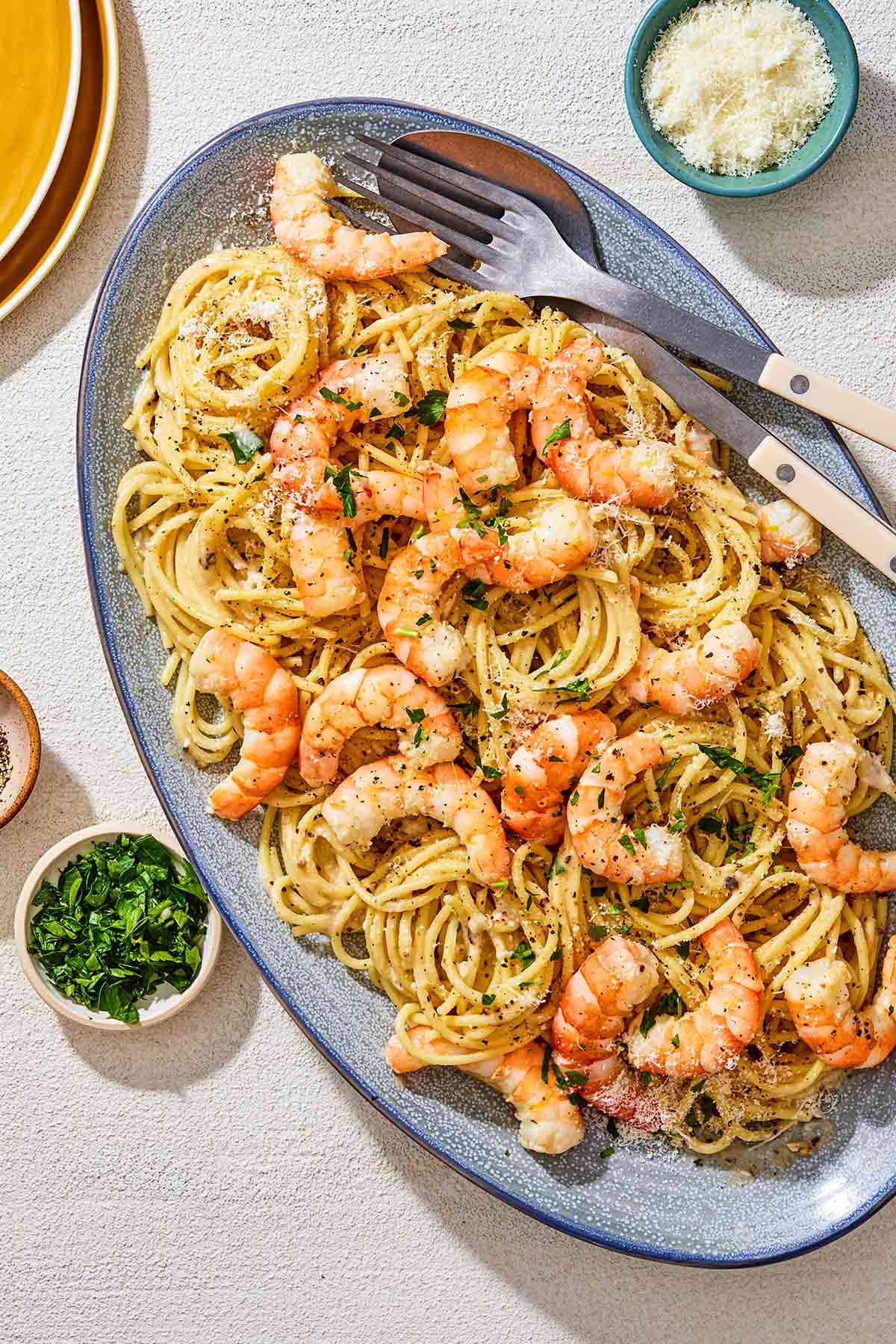 An overhead photo of shrimp cacio e pepe on a serving platter with serving utensils. Next to this is small bowls of pecorino romano cheese and parsley.