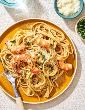 An overhead photo of a plate of cacio e pepe with a fork. Next to this is a glass of water and small bowls of grated pecorino romano cheese and parsley.