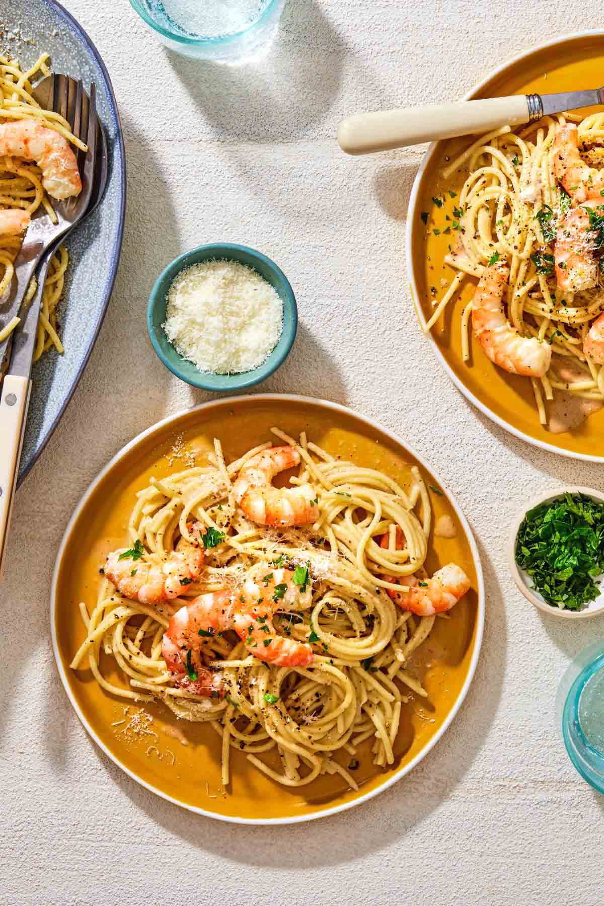An overhead photo of two plates of cacio e pepe, one with a fork. Next to these is a platter with the rest of the shrimp cacio e pepe and serving utensils, small bowls of grated pecorino romano cheese and parsley, and two glasses of water.