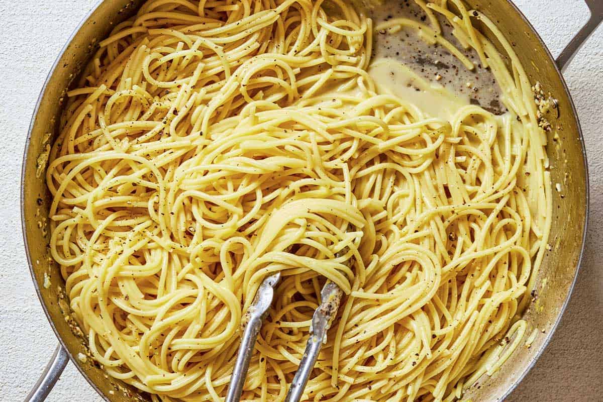 Cooked spaghetti being mixed with the lemon garlic sauce in a skillet with tongs.