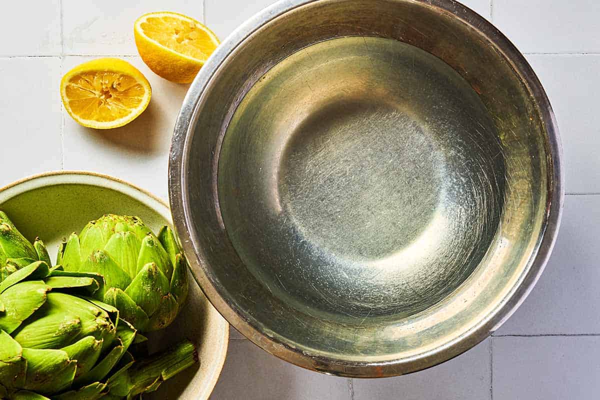 Four globe artichokes in a bowl next to a bowl of water combined with lemon juice, and 2 juiced lemon halves.