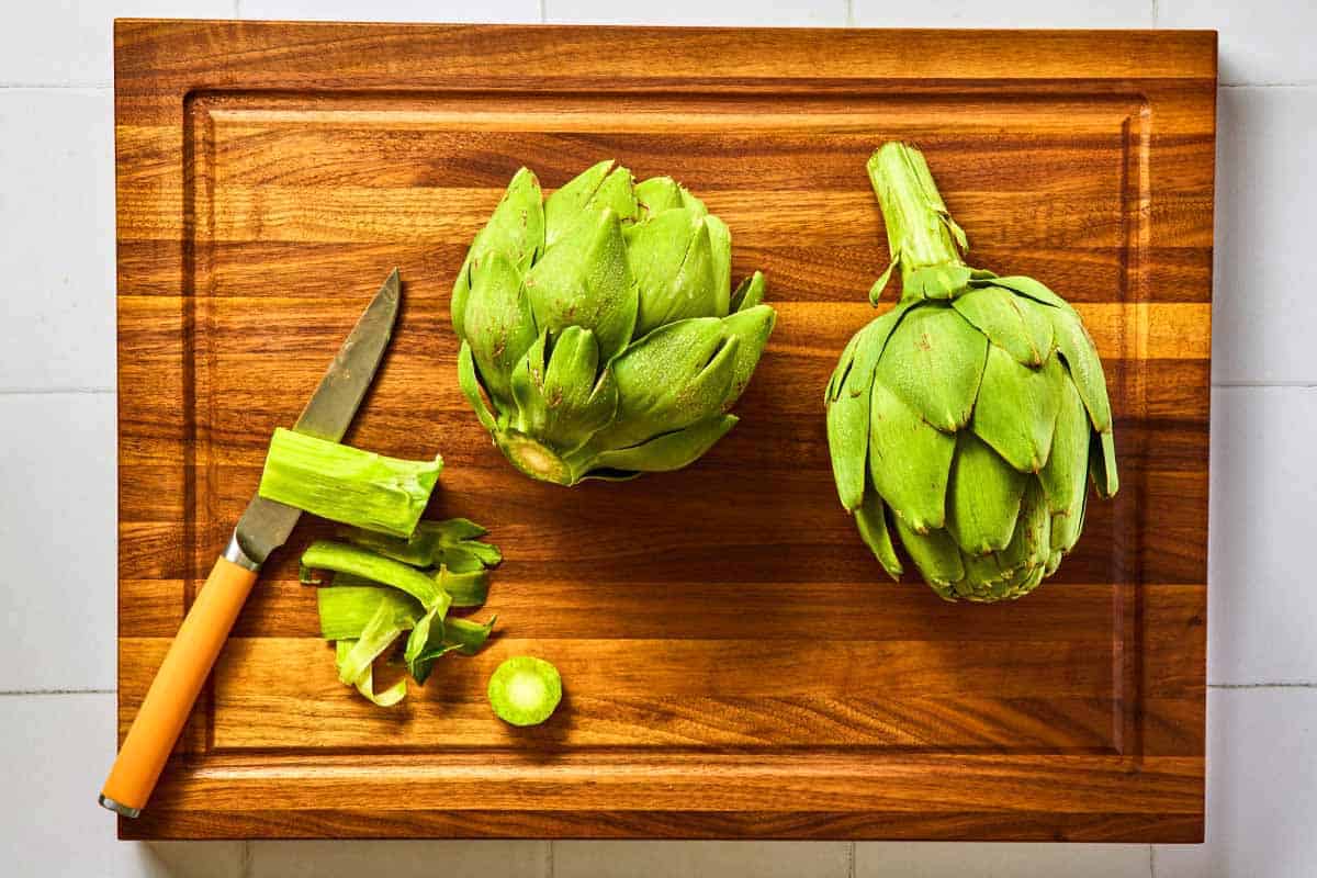 Two globe artichokes on a cutting board, one with it's stem removed with a knife.