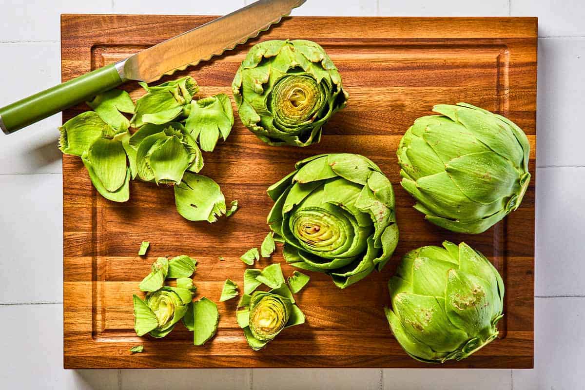 4 globe artichokes with their stems removed on a cutting board in the process of being trimmed with a knife.