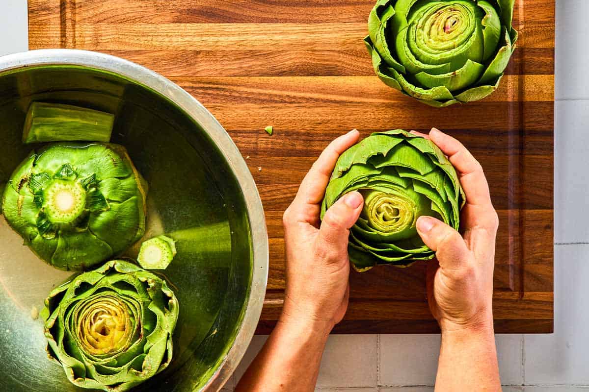 An overhead photo of 2 artichokes in a bowl of lemon water on a cutting board. Also on the cutting board are 2 more artichokes, one with it's leaves being loosened.