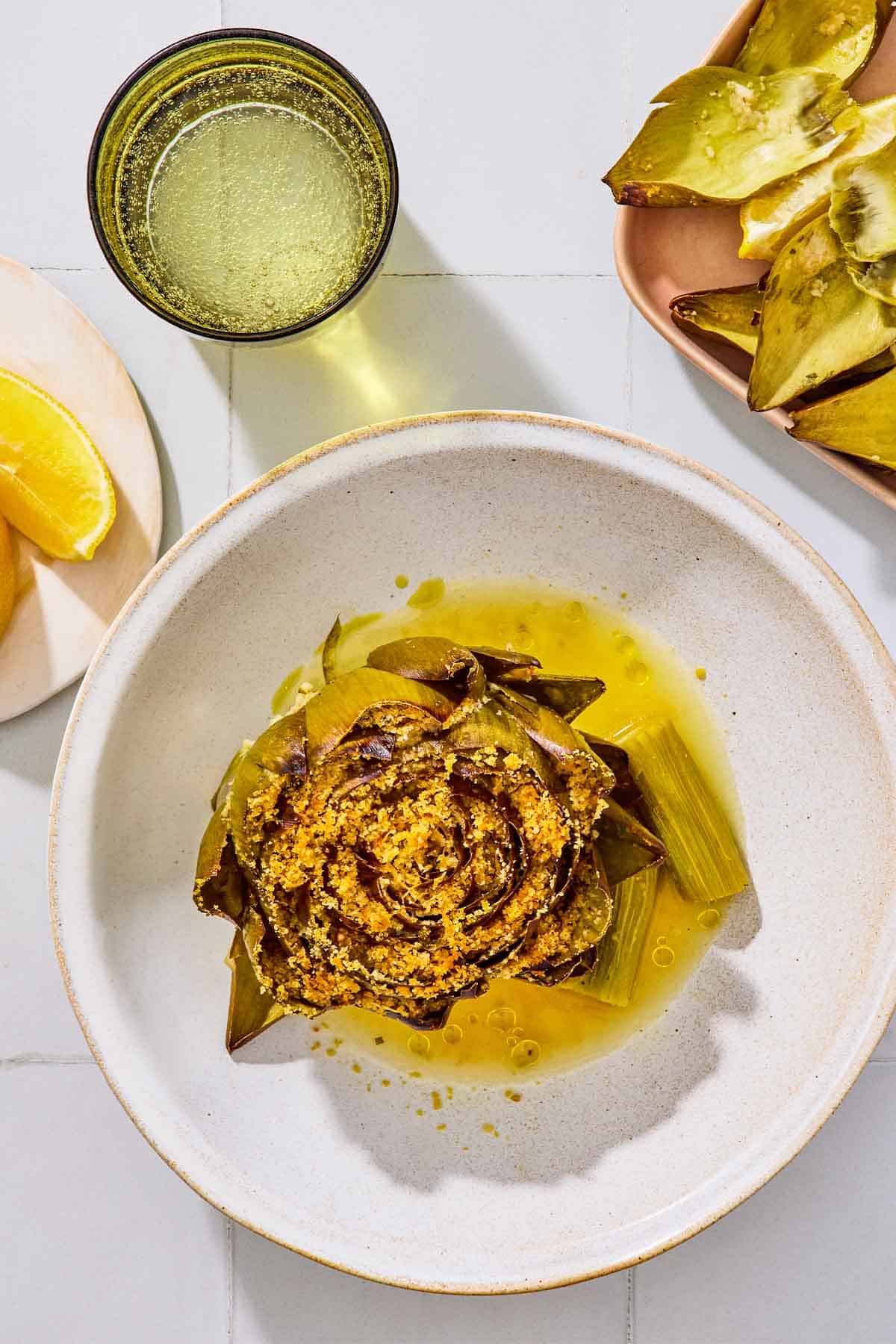 An overhead photo of a cooked stuffed artichoke in a bowl. Next to this is a plate of lemon wedges, a glass of water, and a tray of trimmed artichoke leaves.