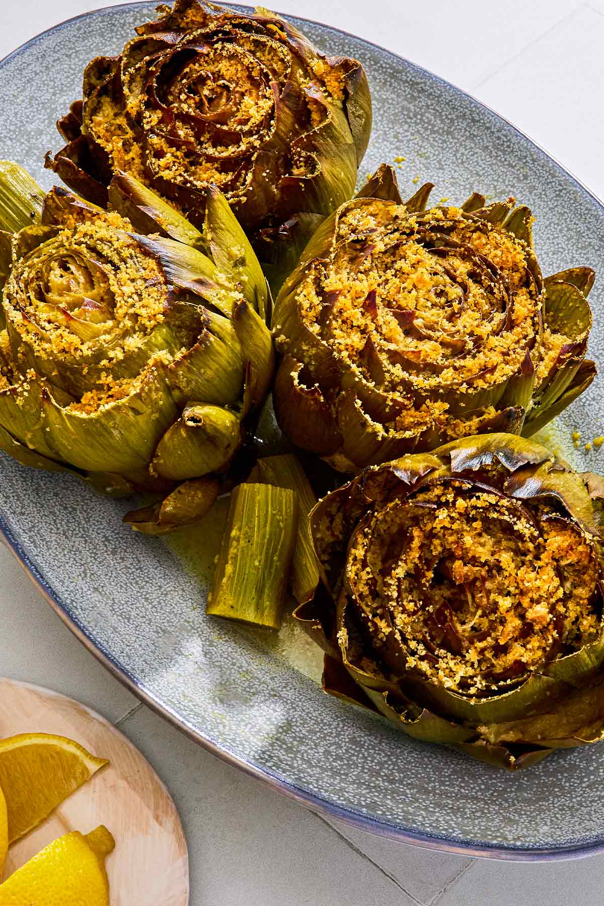 An overhead photo of four cooked stuffed artichokes on a serving patter with their stems. Next to this is a plate of lemon wedges.