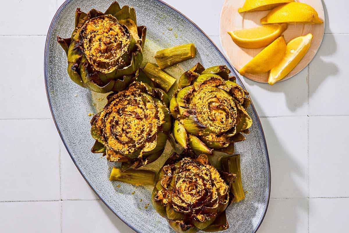 An overhead photo of four cooked stuffed artichokes on a serving patter with their stems. Next to this is a plate of lemon wedges.