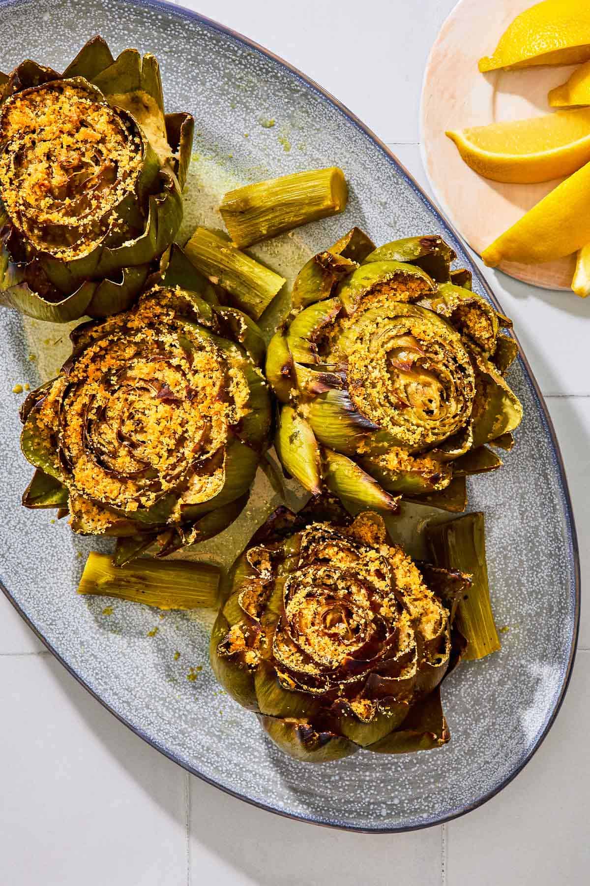 An overhead photo of four cooked stuffed artichokes on a serving patter with their stems. Next to this is a plate of lemon wedges.