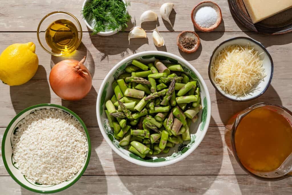 Ingredients for asparagus risotto including asparagus, arborio rice, chicken stock, olive oil, onion, salt, pepper, garlic, lemon, parmesan cheese, and dill.