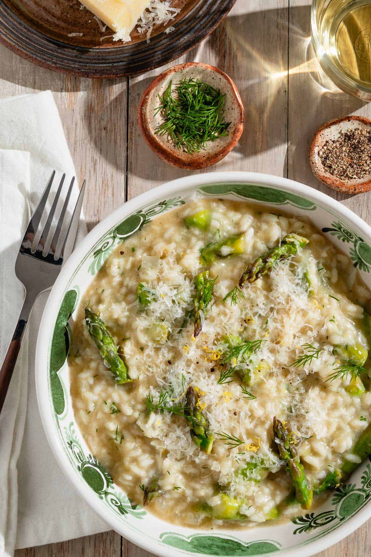 An overhead photo of a serving of asparagus in a bowl next to a fork on a cloth napkin and small bowls of dill and pepper and a glass of wine.