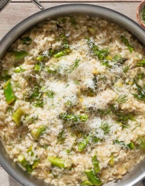 An overhead photo of asparagus risotto topped with parmesan and dill in a skillet. Next to this are small bowls of dill and pepper, a plate with a block of parmesan cheese, and a plate with lemon wedges.