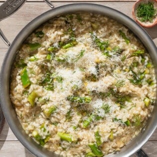 An overhead photo of asparagus risotto topped with parmesan and dill in a skillet. Next to this are small bowls of dill and pepper, a plate with a block of parmesan cheese, and a plate with lemon wedges.