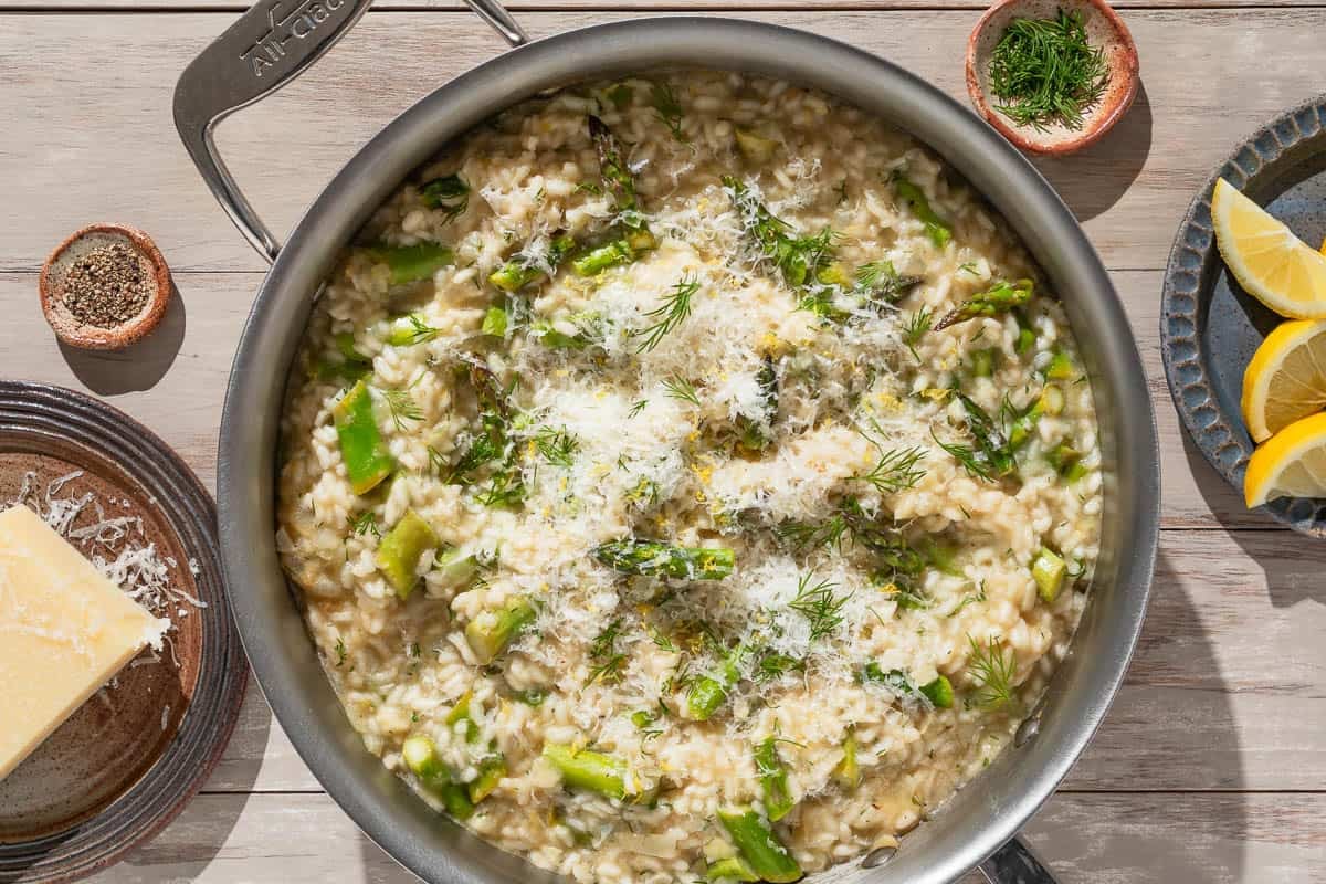 An overhead photo of asparagus risotto topped with parmesan and dill in a skillet. Next to this are small bowls of dill and pepper, a plate with a block of parmesan cheese, and a plate with lemon wedges.