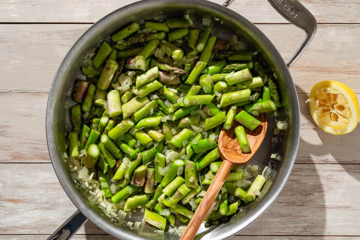 An overhead photo of asparagus, diced onion, garlic, salt, nutmeg and pepper being sauteed in a skillet with a wooden spoon. Next to this is half of a juiced lemon.