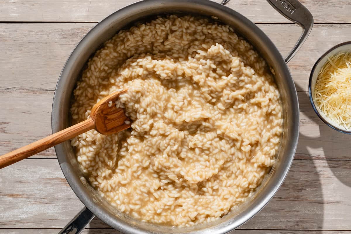 An overhead photo risotto cooking in a skillet with a wooden spoon. Next to this is a bowl of shredded parmesan cheese.