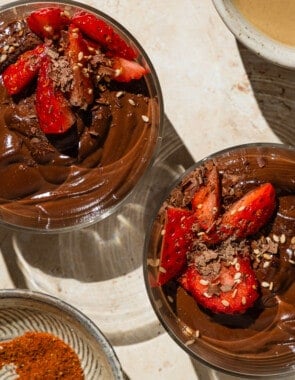 An overhead photo of 2 bowls of vegan chocolate pudding garnished with sliced fresh strawberries. Next to these are bowls of tahini and baharat spice blend, as well as a plate with more strawberries.