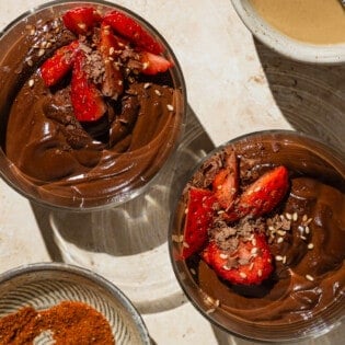 An overhead photo of 2 bowls of vegan chocolate pudding garnished with sliced fresh strawberries. Next to these are bowls of tahini and baharat spice blend, as well as a plate with more strawberries.