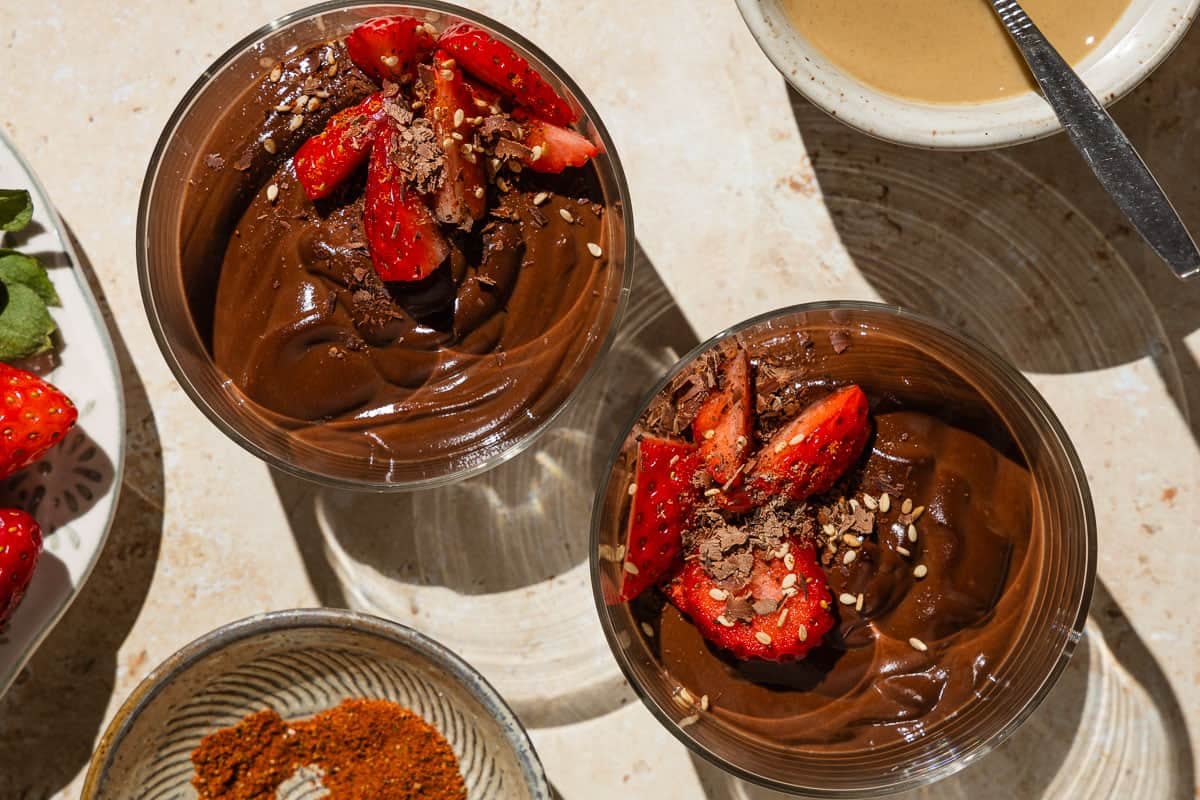 An overhead photo of 2 bowls of vegan chocolate pudding garnished with sliced fresh strawberries. Next to these are bowls of tahini and baharat spice blend, as well as a plate with more strawberries.